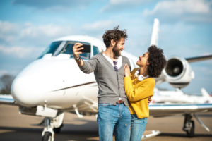 Rich young couple taking a selfie on a mobile phone in front of a private jet parked on an airport tarmac
